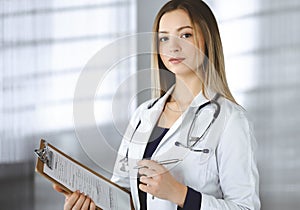 Young woman-doctor is making some notes using a clipboard, while standing in her cabinet in a clinic. Portrait of