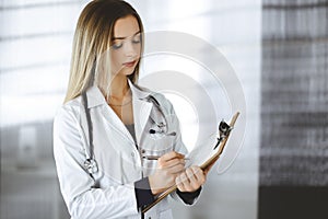 Young woman-doctor is making some notes using a clipboard, while standing in her cabinet in a clinic. Portrait of