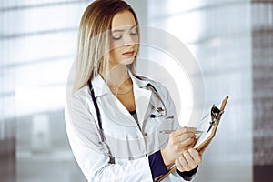 Young woman-doctor is making some notes using a clipboard, while standing in her cabinet in a clinic. Portrait of