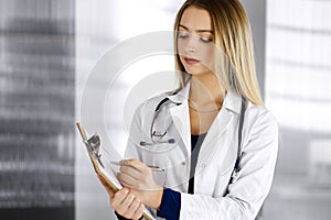 Young woman-doctor is making some notes using a clipboard, while standing in her cabinet in a clinic. Portrait of