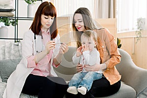 Young woman doctor measuring temperature of baby girl kid. Doctor taking baby`s temperature