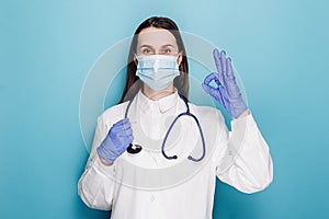 Young woman doctor in medical mask and white coat, rubber gloves, ensure everything alright, show okay gesture