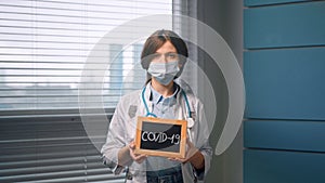 Young woman doctor in white medical coat and face mask holds black covid 19 nameplate and looks straight