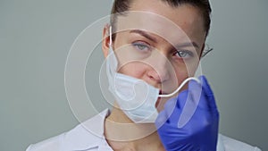 A young woman doctor in a white robe and gloves puts on a protective mask on her face.