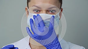 A young woman doctor in a white robe and gloves puts on a protective mask on her face.