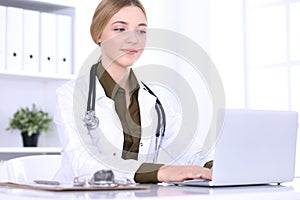 Young woman doctor at work while typing on laptop computer in hospital office. Physician controls medication history