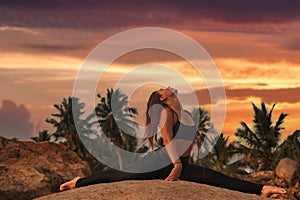 Young woman does yoga position splits or twine for healthy lifestyle on tropical coast ocean outdoors on sunset