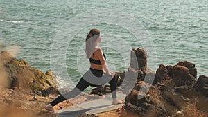 young woman is doing Ashva Sanchalanasana on a seaside rocky shore