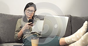 Young woman doing calculations in smartphone checking report or plan on paper while working on laptop at home.