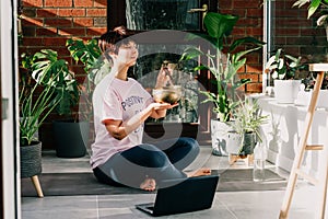 Young woman doing exercise yoga, playing on tibetan singing bowl with its mallet in cozy modern loft style room full of