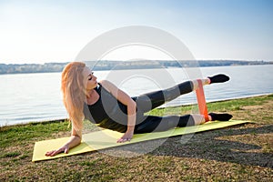 Young woman doing exercises with  resistance band outdoors near the lake at daytime. Healthy lifestyle