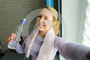 Young woman doing selfies after workout. Smiling attractive fitness girl with towel and bottle of water after training. Sport at