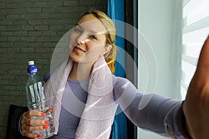 Young woman doing selfies after workout. Smiling attractive fitness girl with towel and bottle of water after training. Sport at