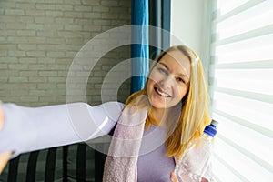 Young woman doing selfies after workout. Smiling attractive fitness girl with towel and bottle of water after training. Sport at