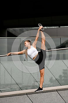 Young woman doing splits stretching, raising arm with leg near glass railing