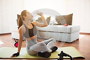 Young woman doing sport workout in room during quarantine. Rest after exercise. Girl sit on mat and drink water from