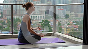 Young woman doing yoga exercises on her balcony in multi-storey building with a view on a downtown with skycrappers