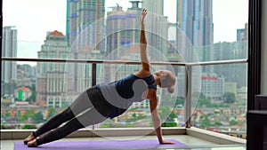 Young woman doing yoga exercises on her balcony in multi-storey building with a view on a downtown with skycrappers