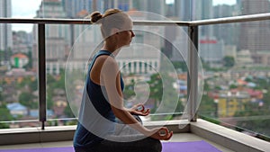 Young woman doing yoga exercises on her balcony in multi-storey building with a view on a downtown with skycrappers