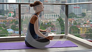 Young woman doing yoga exercises on her balcony in multi-storey building with a view on a downtown with skycrappers