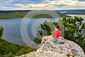 Young woman is doing yoga exercises on the rock above beautiful river.