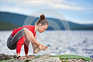 Young woman doing yoga exercises on stone near big river against beautiful landscape.