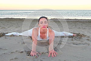 Young woman is doing yoga sitting on a cross twine on the sandy beach at sunrise.