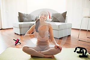 Young woman doing yoga workout in room during quarantine. Cheerful positive girl wear santa claus hat and smile. Show