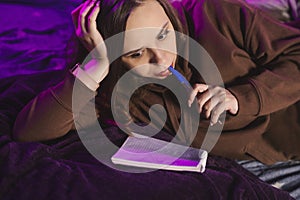 Young woman drawing in notepad, lying in bed at night. Close up. Relaxed female writing in copybook before going to bed