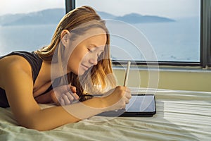 Young woman draws on a tablet against the background of a window with a sea view. Designer, sketching