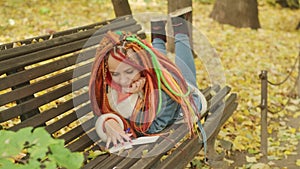 Young woman with dreadlocks drawing, writing in notepad, lying on park bench in golden autumn.