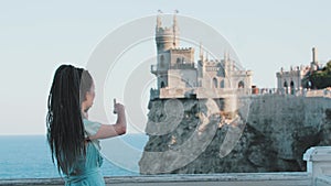 A young woman with dreadlocks stands on a background of sea and a castle on the edge cliff - talking on the phone about