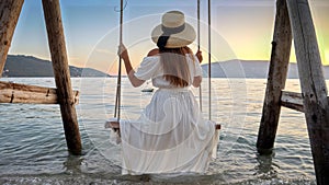 A young woman in a dress gently sways on a swing, her feet playfully touching the sea& x27;s surface, creating splashes