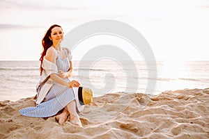 Young woman in a dress holding straw hat and sitting alone on empty sand beach at sunset sea shoreand smiling. Girl looking to the