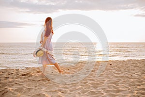 Young woman in a dress holding straw hat and walking alone on empty sand beach at sunset sea shoreand smiling. Single or divorsed
