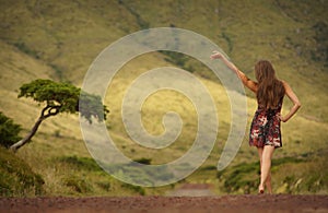 Young woman in dress with raised hand walking along country road. View from behind. Good bye hand raised in the air
