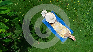 Young woman in dress reads book lying on green grass in a tropical garden on sunny day.