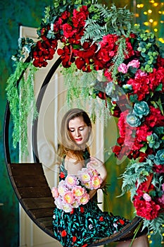 A young woman in a dress sits on a swing with red flowers against a garland of yellow bokeh lights. Holding a bouquet of