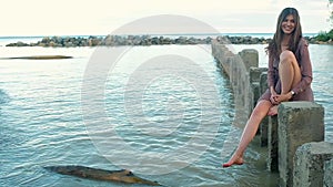 Young woman in dress, sitting on stone pillars, standing in sea making splashes.