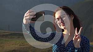 A young woman in a dress stands in the mountains and takes a selfie on a smartphone. The girl travels in the Caucasian