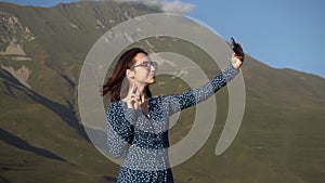 A young woman in a dress stands in the mountains and takes a selfie on a smartphone.