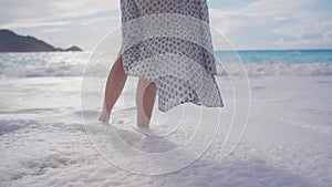 A young woman in a dress stands on the seashore on the beach. The sea washes her feet with foam.