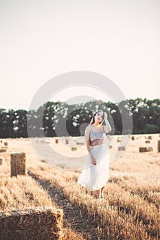 Young woman in dress walking in evening in field with hay bales, beautiful romantic girl with long hair outdoors in field