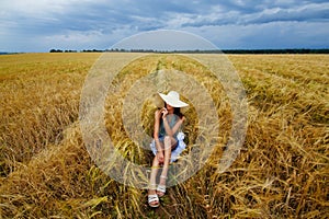 A young woman in a dress and a wide hat, who is sitting in a wheat field before a thunderstorm. Rural landscape in summer