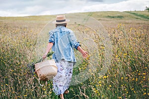 Young Woman dressed jeans jacket and light summer dress walking by the high green grass meadow with basket and wildflowers bouquet