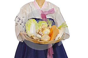 Young woman dressed in a Korean-style white hanbok holds fresh vegetables, white radish, radishes, carrots in a wooden tray, essen