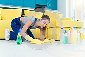 Young woman dressed in special clothes stands on her knees and cleaning the wooden parquet with detergents and rag on