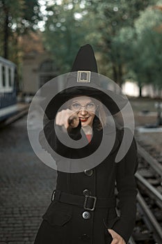 Young woman dressed in witchs hat, black coat and glasses with magical wand in hands. Smiling girl. Vertical frame