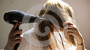 A young woman dries her hair with a hairdryer  on a beige wall background. Art. Young blond woman in a white