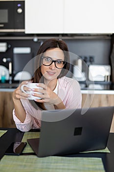 Young woman drink tea working from home using laptop in kitchen